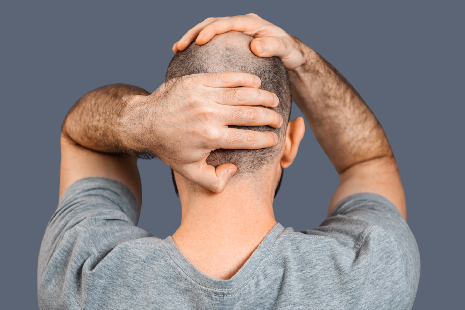 Ein Mann von hinten fotografiert, der seine Hände auf seinen Kopf legt. Er wirkt unsicher und verzweifelt. Das Bild ist in melancholischen Grautönen gehalten, sei T-Shirt sowie der Hintergrund sind grau. 