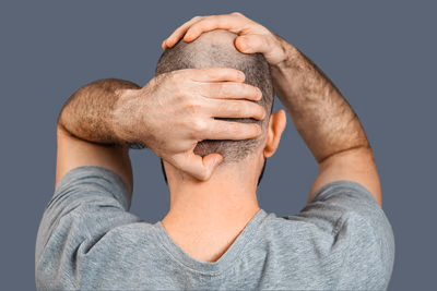 Ein Mann von hinten fotografiert, der seine Hände auf seinen Kopf legt. Er wirkt unsicher und verzweifelt. Das Bild ist in melancholischen Grautönen gehalten, sei T-Shirt sowie der Hintergrund sind grau.