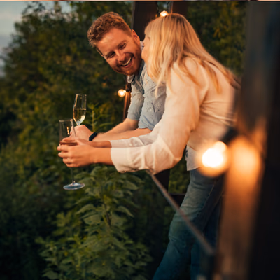 Ein Mann, der eine Escort Lady gebucht hat, steht mit ihr auf einem Balkon oder einer Terrasse im Grünen, umgeben von sanftem Abendlicht und warmen Lichterketten. Beide lachen herzlich miteinander. Die Frau flüstert dem Mann etwas ins Ohr oder küsst ihn liebevoll auf die Wange, während er ein Glas Champagner in der Hand hält. Sie tragen lässige, helle Kleidung – die Stimmung ist intim, sommerlich und ausgelassen. Im Hintergrund sind Bäume und Natur zu sehen, was dem Moment eine entspannte, romantische Atmosphäre verleiht.