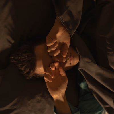 Ein junger Mann liegt in einem Bett mit dunkler Bettwäsche. Er scheint gerade aufzuwachen oder sich gegen das Licht zu schützen – seine Hände sind vor sein Gesicht gehoben, als wolle er die Augen bedecken. Die Szene ist in warmes, weiches Licht getaucht, das eine ruhige und intime Atmosphäre erzeugt. Der Mann trägt ein türkisfarbenes Oberteil, das sich leicht von der dunklen Umgebung abhebt. Sein Gesicht ist nur teilweise sichtbar, da es zum Teil von seinen Händen und der Bettdecke verdeckt wird.